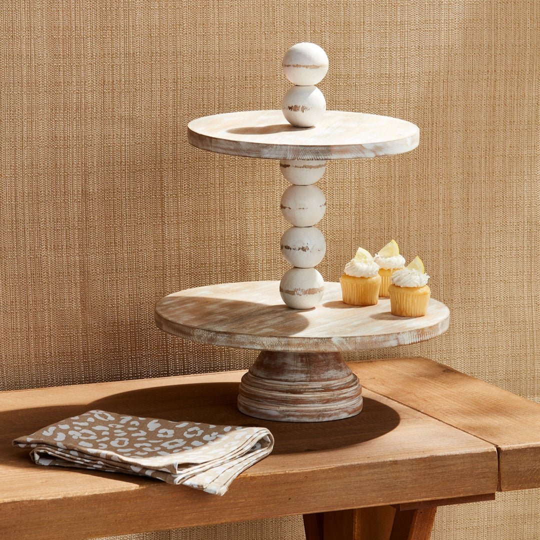Whitewashed wooden two-tiered cupcake stand with a decorative stacked bead pillar displays lemon-topped treats on a wooden table beside a leopard-print cloth napkin against a woven tan backdrop