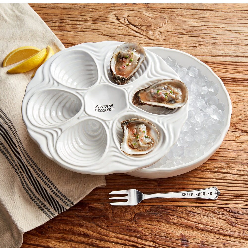 Ceramic Mud Pie oyster serving set displays raw oysters on a scalloped white platter with a central Awww shucks inscription, resting on a bed of crushed ice alongside yellow lemon wedges and a silver fork engraved with SHARP SHOOTER on a rustic wooden table