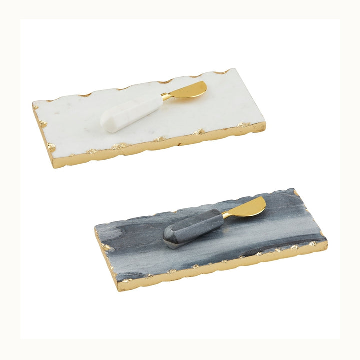 Handcrafted marble and gold-leaf metal rectangular cheese board sets featuring a white and a charcoal grey platter with organic scalloped edges, shimmering gold-foil borders, and matching spreaders with faceted stone handles displayed on a white background