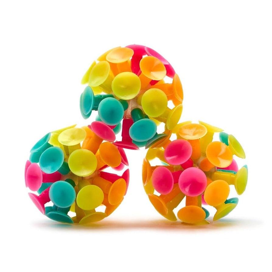 Studio photograph of three multicolored suction cup toy balls stacked in a pyramid on a white background featuring vibrant neon shades of pink, lime green, and orange arranged in a spherical lattice structure