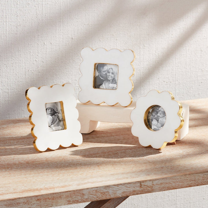 Hand-painted ceramic picture frames by Mud Pie featuring three white frames with scalloped edges and gold-gilded rims displaying black and white family portraits on a rustic wooden surface under soft window light shadows