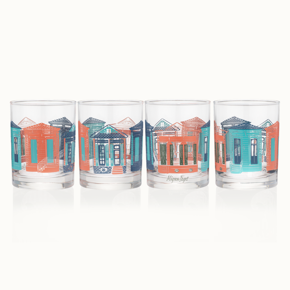 Glassware by Mignon Faget featuring illustrated New Orleans shotgun houses and Creole cottages with vibrant teal and terracotta orange facades, intricate shutter details, and mirror-like reflections on a white background