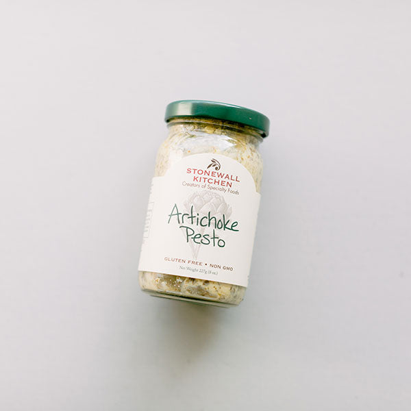 Photograph of a Stonewall Kitchen Artichoke Pesto glass jar on a neutral grey background featuring a dark green lid, hand-written style lettering, and a detailed artichoke illustration on the white label
