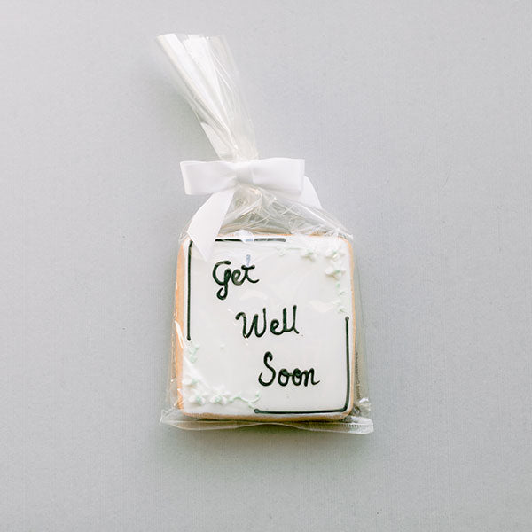 Photograph of a square sugar cookie featuring white royal icing and black cursive Get Well Soon lettering, packaged in a clear cellophane bag with a white satin bow and delicate mint-green floral accents on a neutral gray background