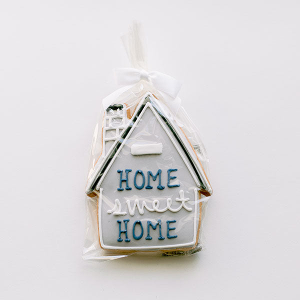 Sugar cookie with royal icing of a house-shaped gift on a white background featuring grey frosting, blue and white lettering reading HOME sweet HOME, and a white chimney detail wrapped in a clear cellophane bag with a white ribbon bow