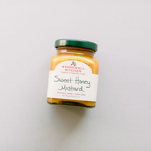 Professional product photograph of a Stonewall Kitchen Sweet Honey Mustard jar centered on a minimalist light gray background featuring a dark green metal lid, textured golden-yellow contents, and a white label with elegant black handwritten script and a red brand logo