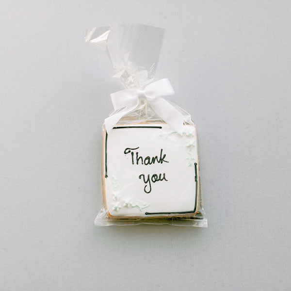 Iced sugar cookie featuring handwritten black cursive Thank you text on white royal icing, accented with delicate white floral designs and minimalist black border lines, wrapped in a clear gift bag with a white satin bow against a neutral grey background