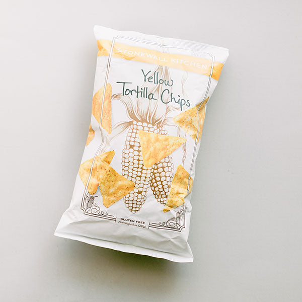 Photograph of a Stonewall Kitchen Yellow Tortilla Chips bag on a neutral grey background featuring a vibrant orange brand label, a detailed black-and-white corn cob sketch, and golden yellow chips depicted across the white packaging