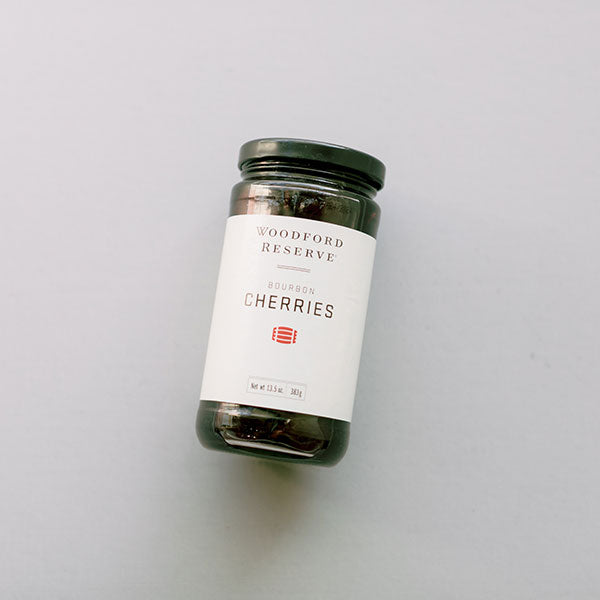 Product photograph of a jar of Woodford Reserve Bourbon Cherries on a solid gray background featuring a white label with a red barrel emblem and dark fruit visible through the tilted glass jar