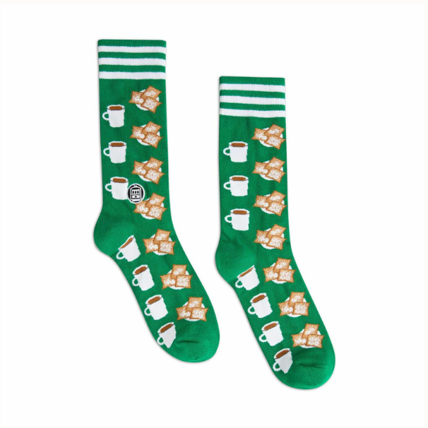 Knitted graphic socks by Bonfolk showcasing a New Orleans theme with a St. Charles streetcar emblem, rows of white cafe au lait mugs and powdered sugar-dusted beignets on a vibrant emerald green fabric with triple white top stripes