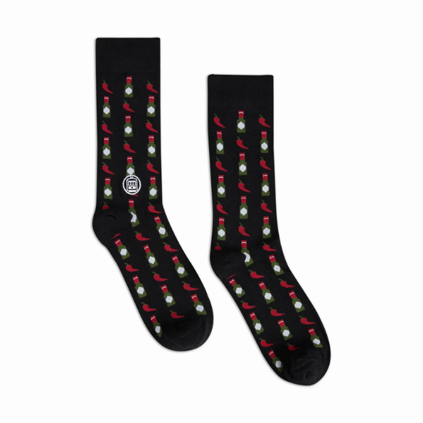 Graphic crew socks by NOLA Couture featuring a black fabric base decorated with a repeating pattern of vibrant red chili peppers, green-capped hot sauce bottles, and the iconic white St. Charles Streetcar emblem from New Orleans