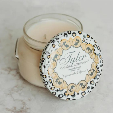 Commercial photograph of a Tyler Candle Company scented jar candle on a white marble surface featuring a decorative leopard-print lid with gold scrollwork leaning against an open glass jar of cream-colored wax