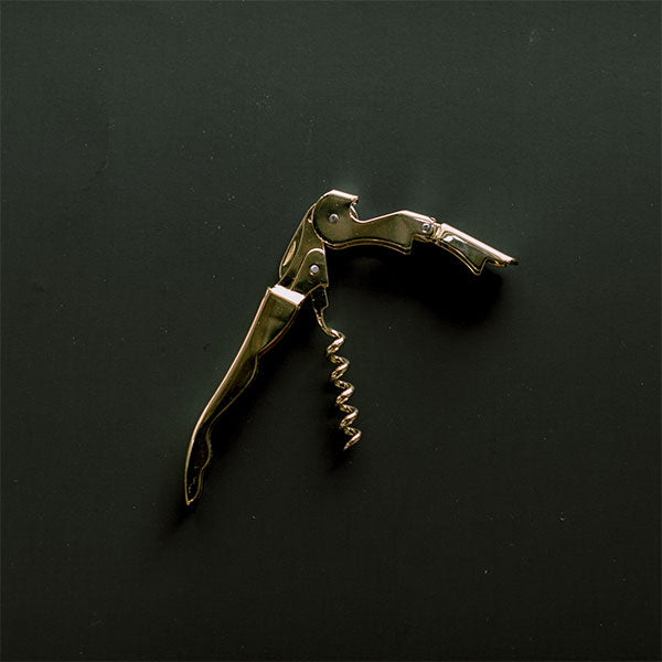 Photograph of a polished gold waiter’s friend corkscrew set against a dark textured background featuring a gleaming metallic surface and a sharp spiraled worm