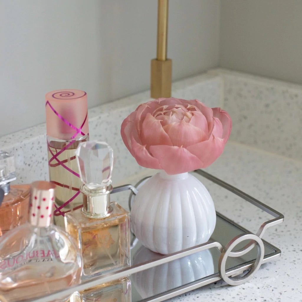 Photograph of a vanity arrangement on a speckled bathroom counter featuring a large pink flower in a white ribbed vase and assorted perfume bottles on a silver mirrored tray including one with a crystal stopper and another with a pink spiral ribbon design