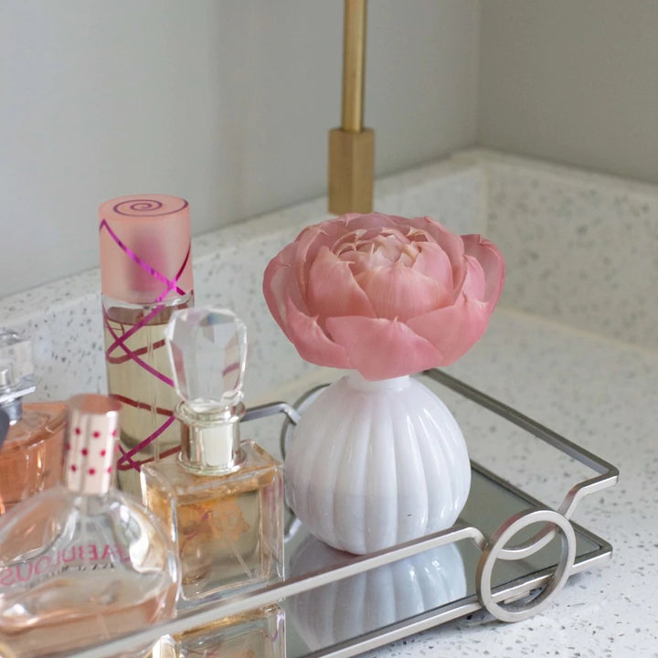 Photograph of a vanity arrangement on a speckled bathroom counter featuring a large pink flower in a white ribbed vase and assorted perfume bottles on a silver mirrored tray including one with a crystal stopper and another with a pink spiral ribbon design