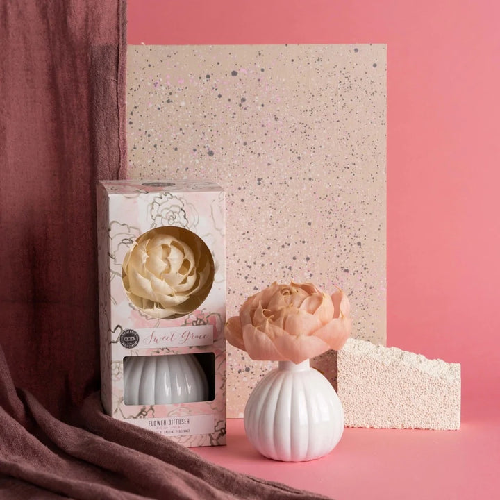 Studio product photograph of a Bridgewater Candle Company Sweet Grace Flower Diffuser featuring a peach-colored sola wood flower in a white fluted ceramic vase against a speckled beige backdrop and draped mauve fabric on a pink surface