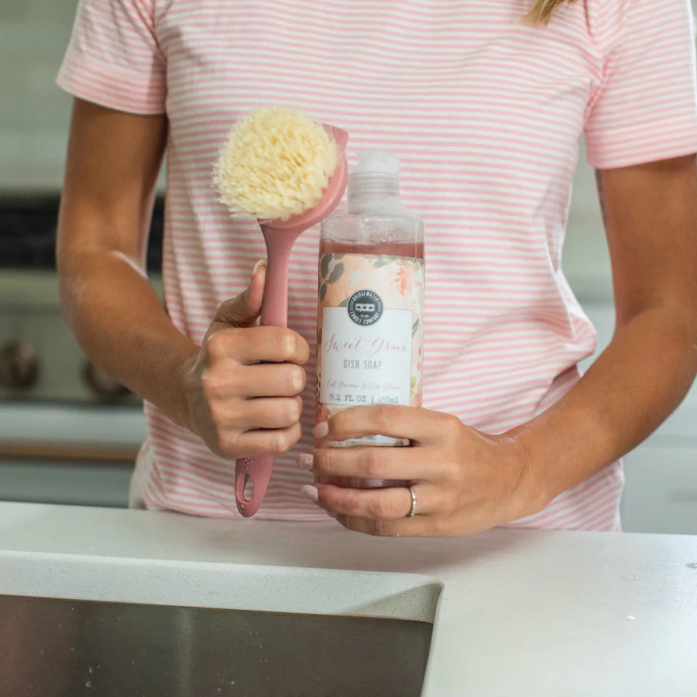 Photograph of a woman wearing a pink and white striped shirt holding a bottle of Sweet Grace dish soap and a matching pink scrub brush with cream bristles in a kitchen sink area