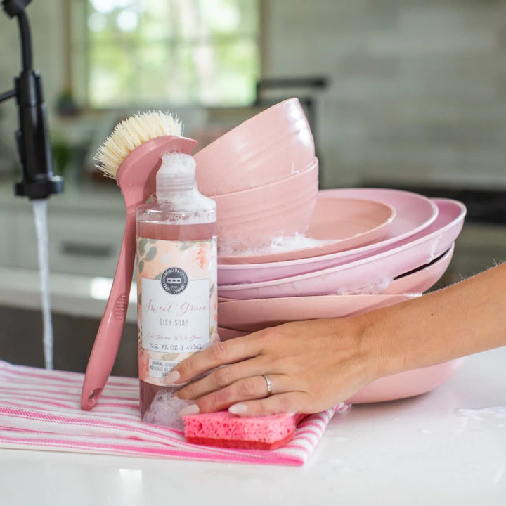 Photograph by Bridgewater Candle Company of Sweet Grace Dish Soap on a white kitchen counter featuring a stack of blush pink dishes, a matching pink scrub brush, and a hand with a wedding ring resting on a pink sponge and striped towel