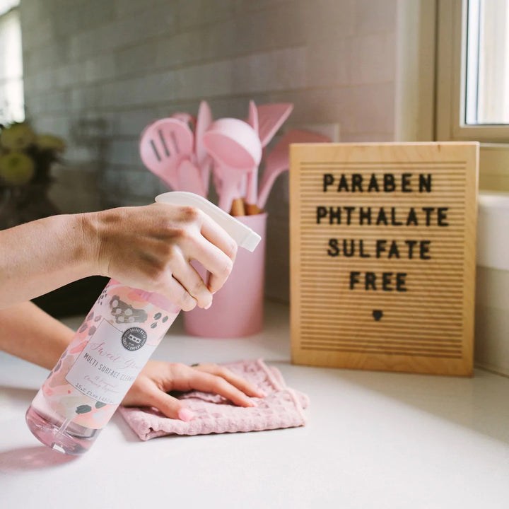 Lifestyle photograph by Bridgewater Candle Company of a person spraying Sweet Grace Multi Surface Cleaner on a white kitchen counter featuring a pink textured cloth and a wooden letter board specifying paraben phthalate and sulfate free ingredients alongside pink cooking utensils