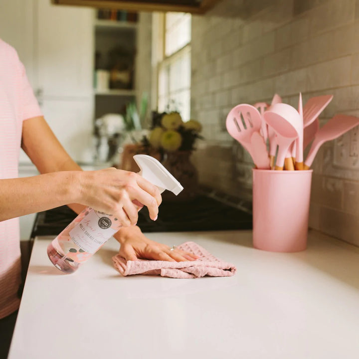 Photograph of a person cleaning a white kitchen countertop with a pink-labeled multi-surface spray bottle, a textured pink cloth, and a pink canister of matching silicone utensils against a white subway tile backsplash