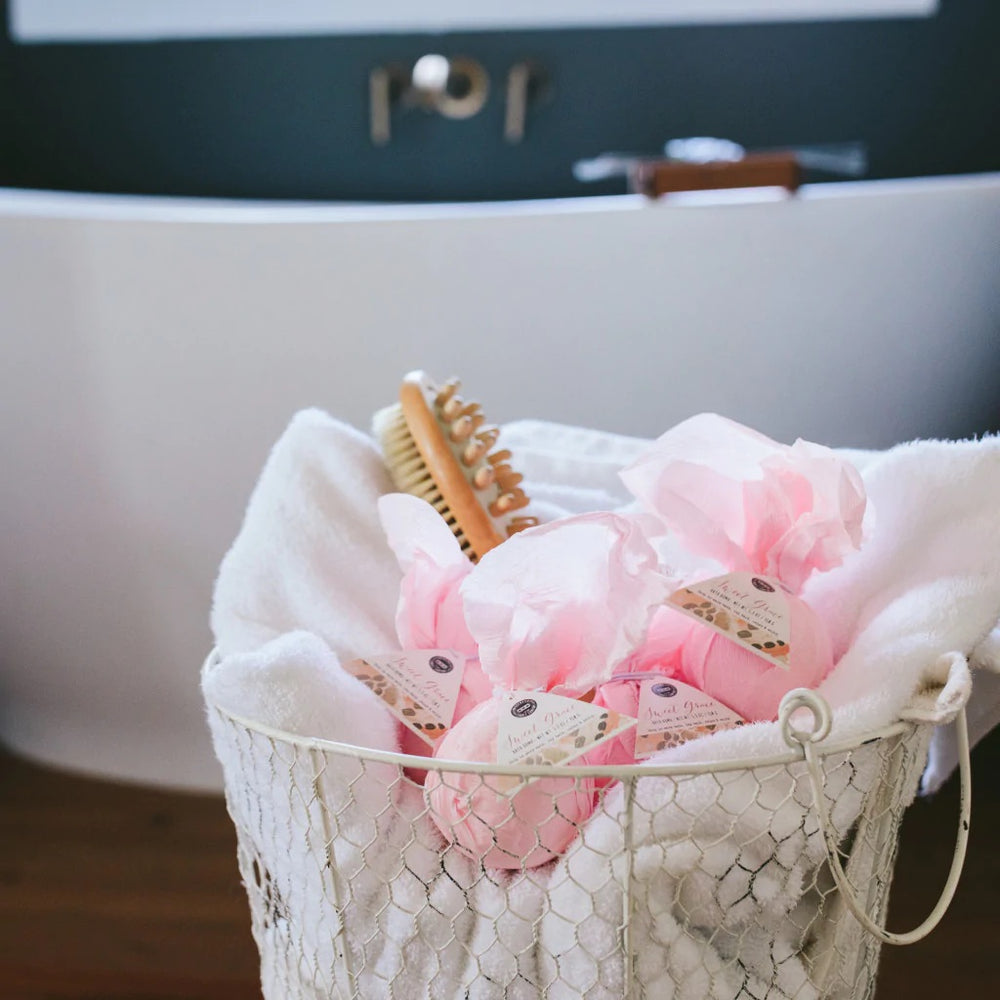 Digital lifestyle photograph of pink Sweet Grace bath bombs and a wooden massage brush in a white wire basket featuring a modern white soaking tub and dark wall-mounted faucet
