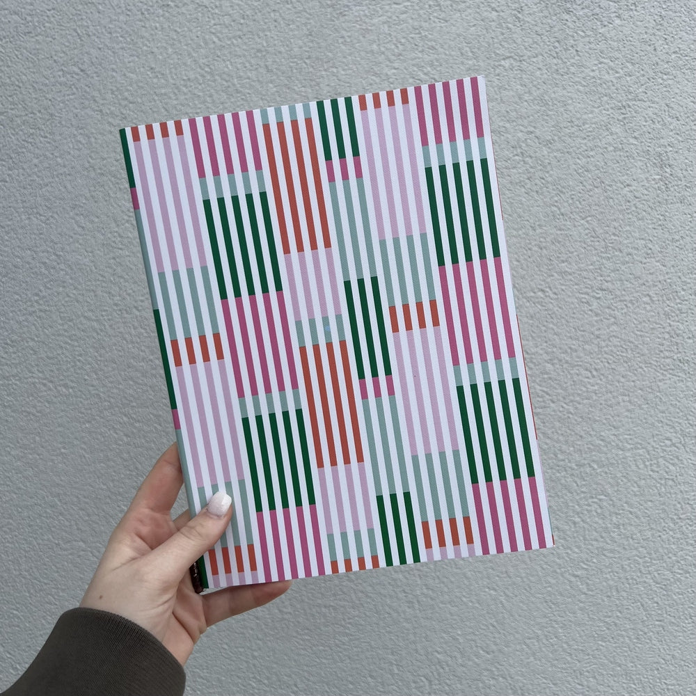 Graphic design notebook featuring a vibrant geometric pattern of forest green, coral orange, and bubblegum pink vertical stripes held by a hand with white nails against a gray textured wall