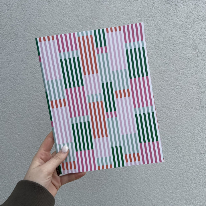 Graphic design notebook featuring a vibrant geometric pattern of forest green, coral orange, and bubblegum pink vertical stripes held by a hand with white nails against a gray textured wall
