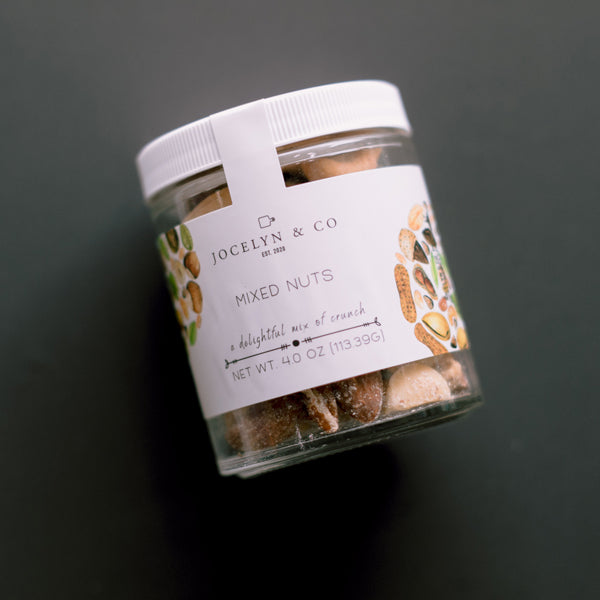 Digital photograph by Jocelyn &amp; Co featuring a clear jar of mixed nuts on a dark background, highlighting a white label with colorful watercolor nut illustrations, minimalist branding, and a white ribbed lid