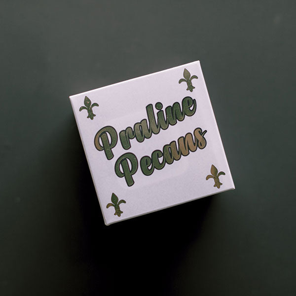 Digital photograph of a New Orleans Praline Pecans box featuring stylized green script and four iconic gold fleur-de-lis symbols set against a dark minimalist background