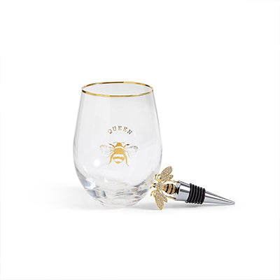 Glass and metal barware featuring a gold-rimmed stemless wine glass with a queen bee graphic and a matching bejeweled bee bottle stopper with a silver conical base on a white background