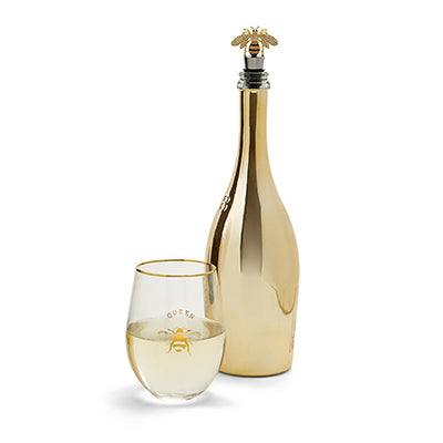 Studio photograph of a gold-toned barware set featuring a metallic champagne bottle with an ornate honeybee stopper and a stemless wine glass displaying the word QUEEN above a golden bee motif on a white background