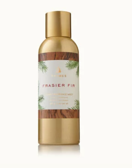 Commercial product photography of a Thymes Frasier Fir Home Fragrance Mist bottle on a white studio background featuring a gold metallic finish, wood-grain label borders, and delicate green pine needle illustrations