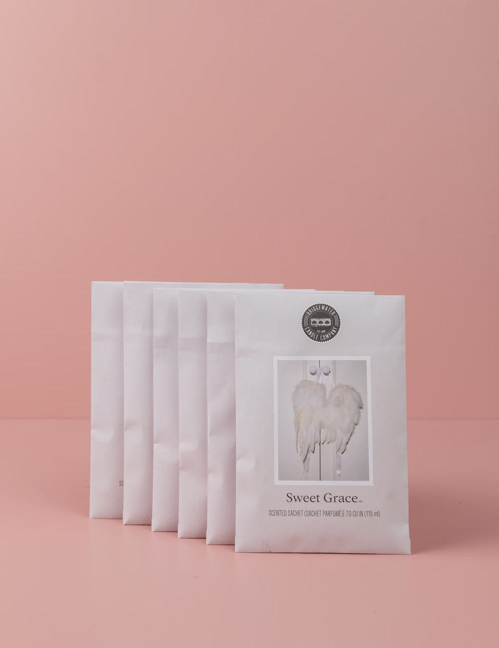 Commercial studio photograph by Bridgewater Candle Company featuring a staggered row of six Sweet Grace scented sachets against a solid pink backdrop showcasing white feathery angel wings and a circular black brand logo