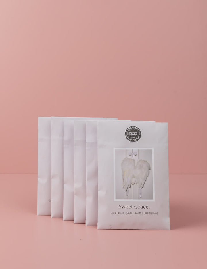 Commercial studio photograph by Bridgewater Candle Company featuring a staggered row of six Sweet Grace scented sachets against a solid pink backdrop showcasing white feathery angel wings and a circular black brand logo