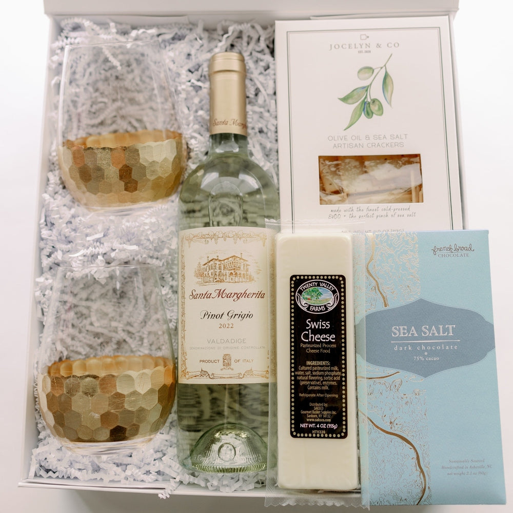 Photograph of a gourmet gift box featuring a bottle of Santa Margherita Pinot Grigio, two stemless wine glasses with gold honeycomb-patterned bases, and a light blue bar of French Broad chocolate resting on white crinkle paper