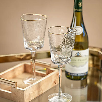 Photograph of two hammered texture wine glasses with gold-rimmed irregular edges displayed on a mirrored surface beside a bottle of white wine and a light wood tray with gold handles