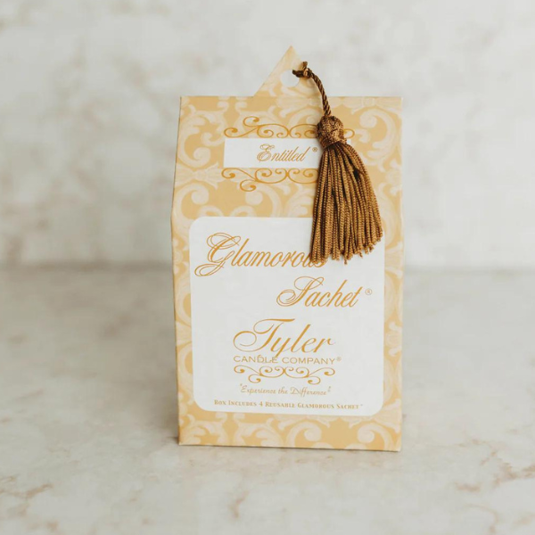 Commercial product photograph by Tyler Candle Company of an Entitled scent Glamorous Sachet box on a marble surface featuring the brand script logo with a golden damask pattern and a decorative bronze tassel