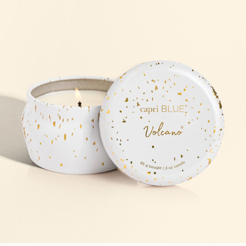 Scented candle by Capri Blue featuring a white tin with metallic gold splatter, a small lit wick, and the iconic Volcano logo on the propped-up lid against a cream background