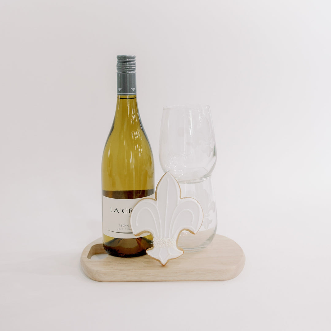 Photograph of a New Orleans inspired gift set featuring a bottle of La Crema Monterey Chardonnay, stacked Mignon Faget stemless glasses with fleur-de-lis etchings, and a white frosted fleur-de-lis sugar cookie on a light wood board