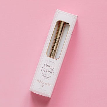 Product photograph for Baublerella featuring the Bling Brush jewelry cleaner on a pink background with a gold patterned pen visible through a window and elegant white packaging with serif typography
