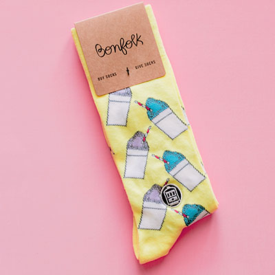 Cotton socks by Bonfolk featuring a New Orleans-inspired repeat pattern of blue and purple snow cones and an iconic streetcar emblem on vibrant yellow fabric against a solid pink background
