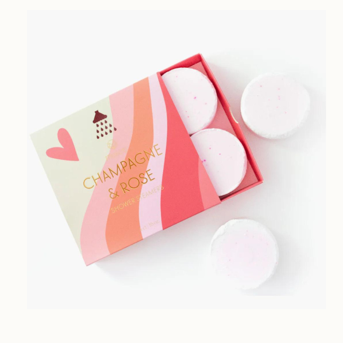 Commercial product photograph of Champagne and Rose Shower Steamers featuring a pink and peach wavy-patterned box with gold foil lettering, a stylized heart icon, and round white tablets with delicate pink speckles on a white background