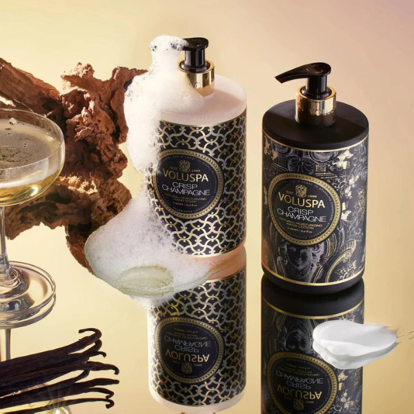 Professional product photograph of Voluspa Crisp Champagne soap and lotion on a gold mirrored surface featuring overflowing white foam, a cream smear, and dark vanilla beans