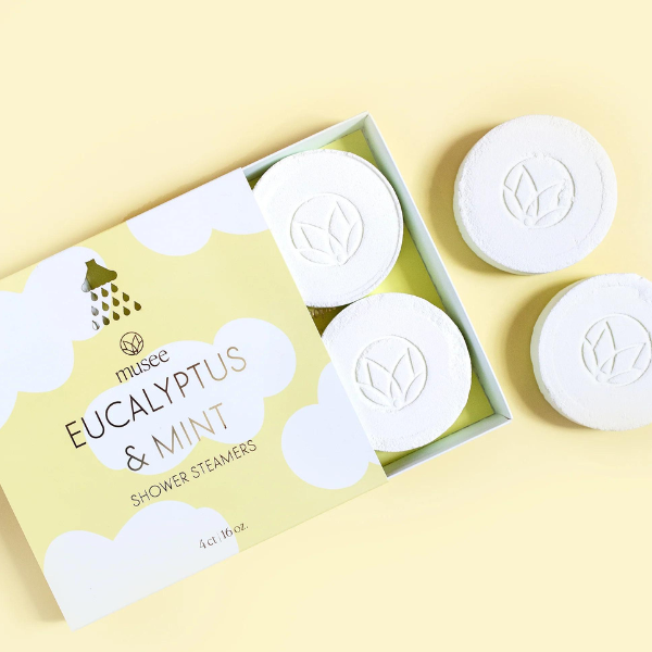 Product photograph by Musee of Eucalyptus and Mint shower steamers in a yellow cloud-patterned box on a pale surface featuring white circular tablets with embossed lotus logos and a metallic gold shower head icon