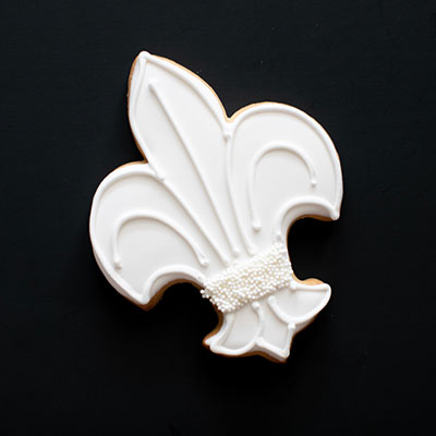Iced sugar cookie of an iconic fleur-de-lis symbol featuring smooth white royal icing, elegant raised scrollwork, and a central band of tiny white nonpareils against a solid black background