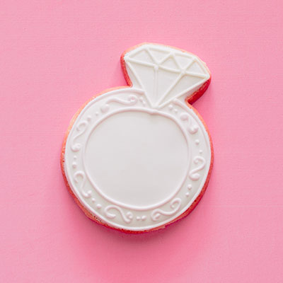 Sugar cookie shaped like a diamond engagement ring centered on a vibrant pink background featuring intricate white royal icing filigree and a geometric faceted diamond design