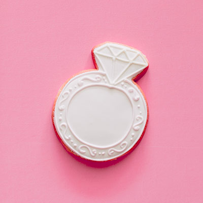 Sugar cookie with royal icing shaped like a diamond engagement ring centered on a vibrant pink background featuring intricate white piped swirls, decorative dots, and a faceted diamond-shaped topper