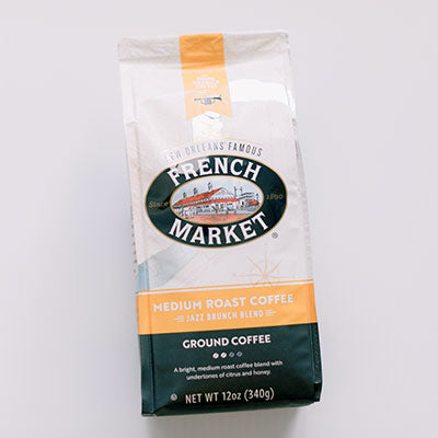 Commercial product photograph of a bag of French Market Jazz Brunch Blend ground coffee from New Orleans featuring an illustration of the historic French Market building with red roofs, bold orange and dark green accents, and a small black trumpet icon