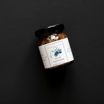 Studio photograph of a Jocelyn & Co two-ounce jar of olive tapenade on a matte black background featuring a white label with a botanical watercolor illustration of blue olives and a glossy black lid