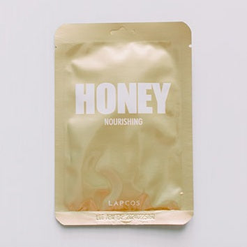 Product photograph by Lapcos of a honey nourishing sheet mask packet centered on a white background featuring a metallic gold foil finish with bold white capital letters and an embossed crimped seal at the bottom